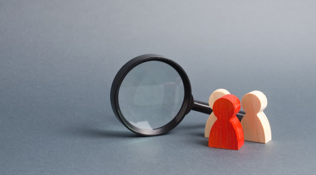 A magnifying glass focusing on two wooden figurines, one painted red and the other in natural wood, set against a plain grey background.