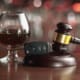 A gavel lies on its side next to a car key and a glass of dark liquor in the background, sitting on a wooden surface with a blurred, colorful backdrop. The scene evokes themes of legal issues and drinking and driving.