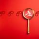 A magnifying glass with a gavel and sound block icon inside is centered on a red background. The shadowy outline of several pairs of handcuffs is visible in a horizontal row behind the magnifying glass.